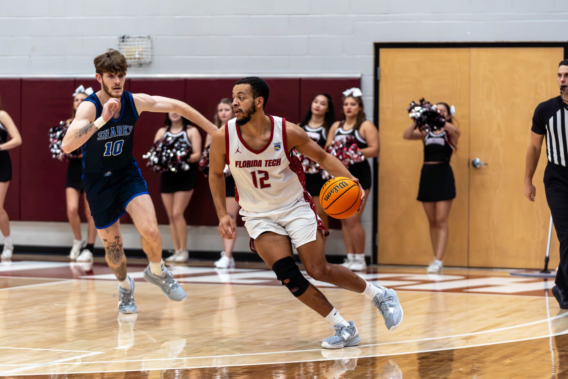 A basketball game at Florida Tech where a player in a white jersey with the number 12 dribbles the ball while being pursued by an opponent in a blue jersey. Cheerleaders and a referee are visible in the background